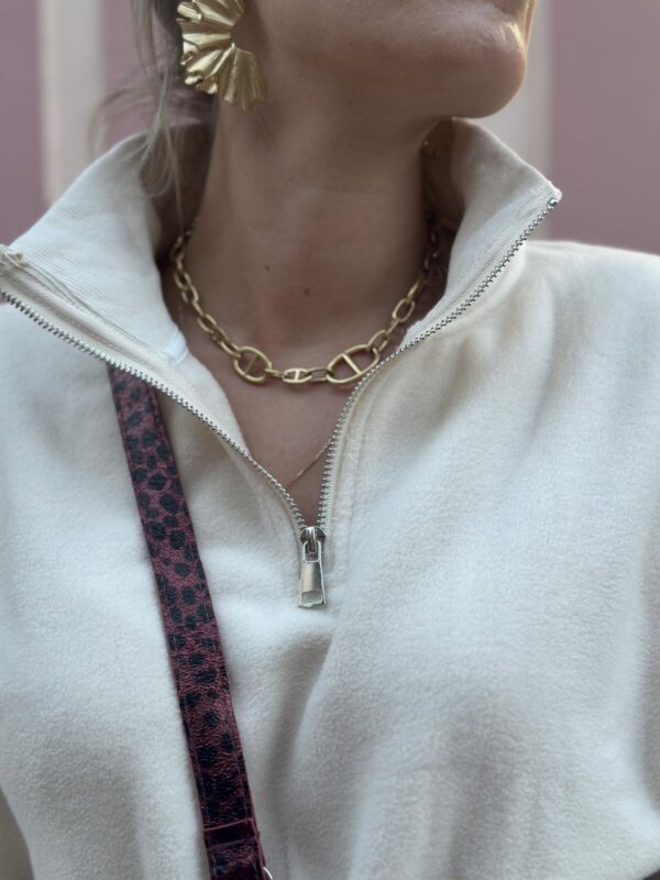 Pull mégane porté avec un zoom rapproché au niveau du col. Porté avec un collier couleur or et des boucles d'oreilles or. La photo est prise sur un fond rose.