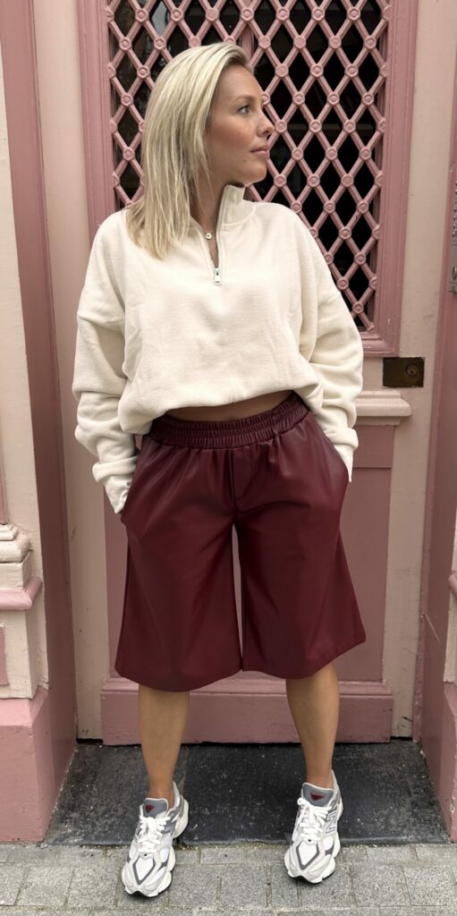 Short MILO bordeaux avec un élastique au niveau de la taille. Un short aspect vintage mais très moderne, un visuel en similicuir. Porté avec un pull a fermeture éclaire et des basket blanche. La photo est prise sur un mur rose.