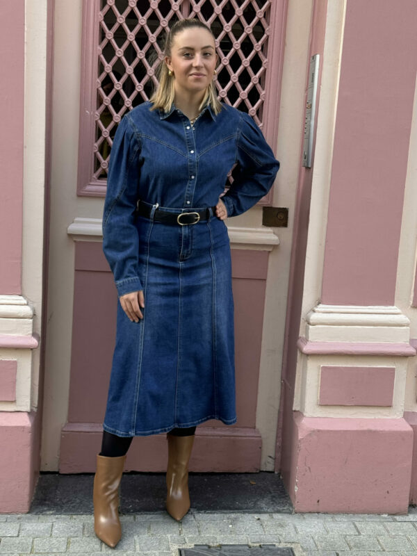 Chemise SALOME couleur bleu jeans, avec des boutons à clipser écru perlé. Porté avec une jupe dans le même coloris que la chemise, une ceinture noire boucle dorée et des bottines camel LOUISE à bout pointu disponible sur le site internet. La photo est prise sur un fond rose